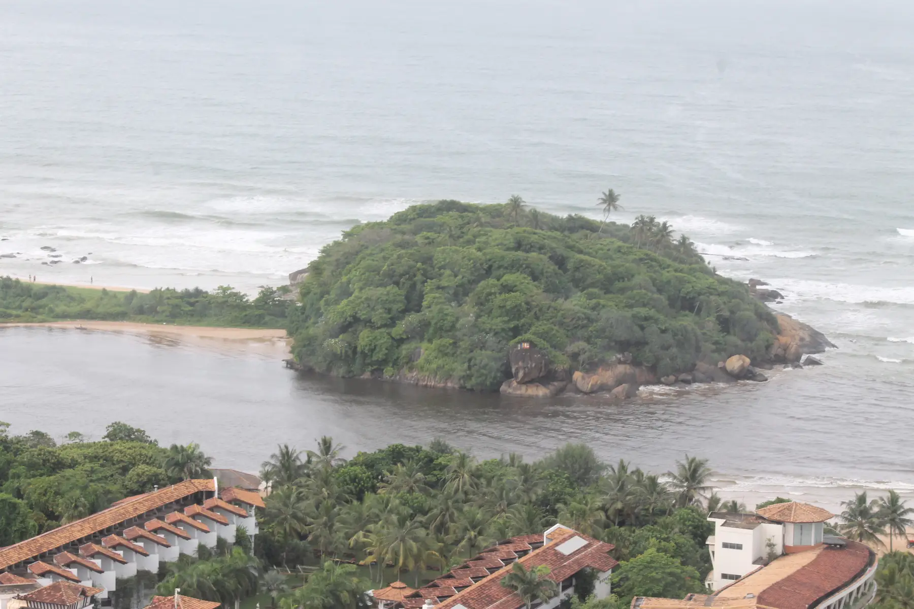 The Bentota river estuary seen from above, with a wooded rock headland, a sand spit and the sea beyond.