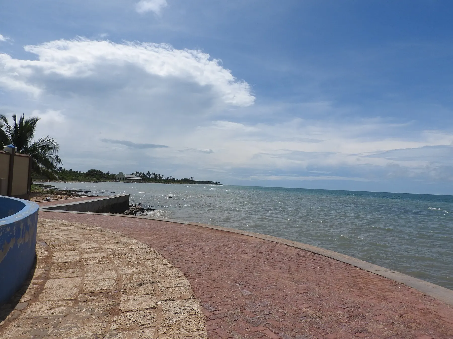 A curved red-brick sea promenade at Keerimalai on the Jaffna peninsula, with calm blue water and white clouds beyond.
