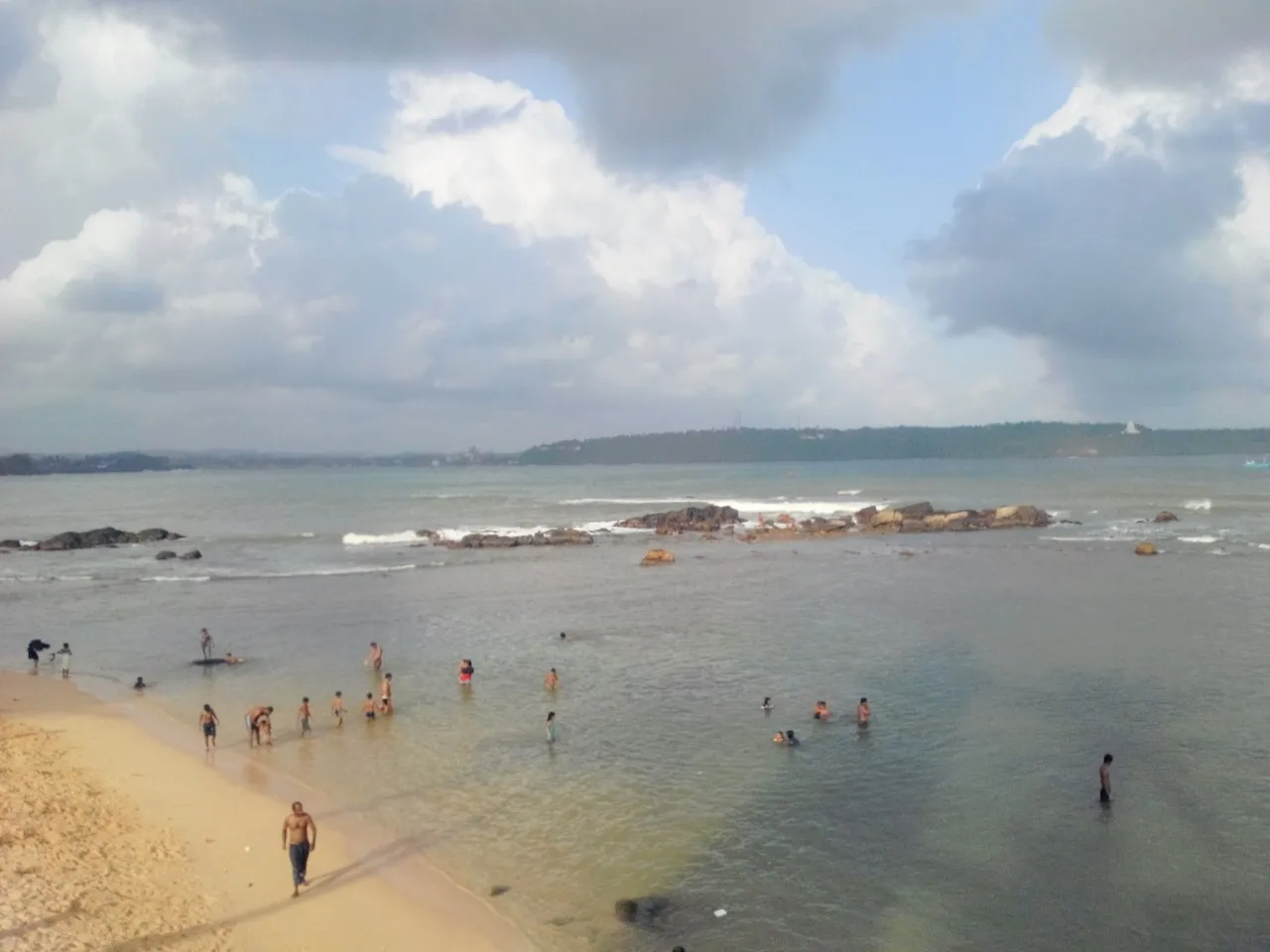 Shallow water at Unawatuna beach with swimmers in the bay, rocks offshore and a wooded headland behind.