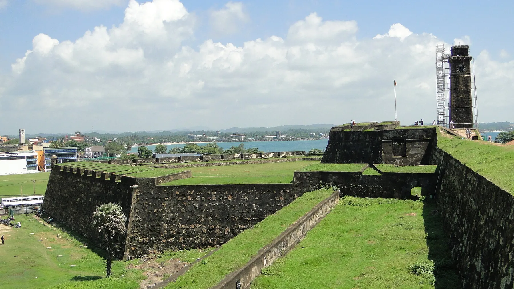 Grassy ramparts and dark stone bastion walls of Galle Fort under a blue sky, with the clock tower behind.