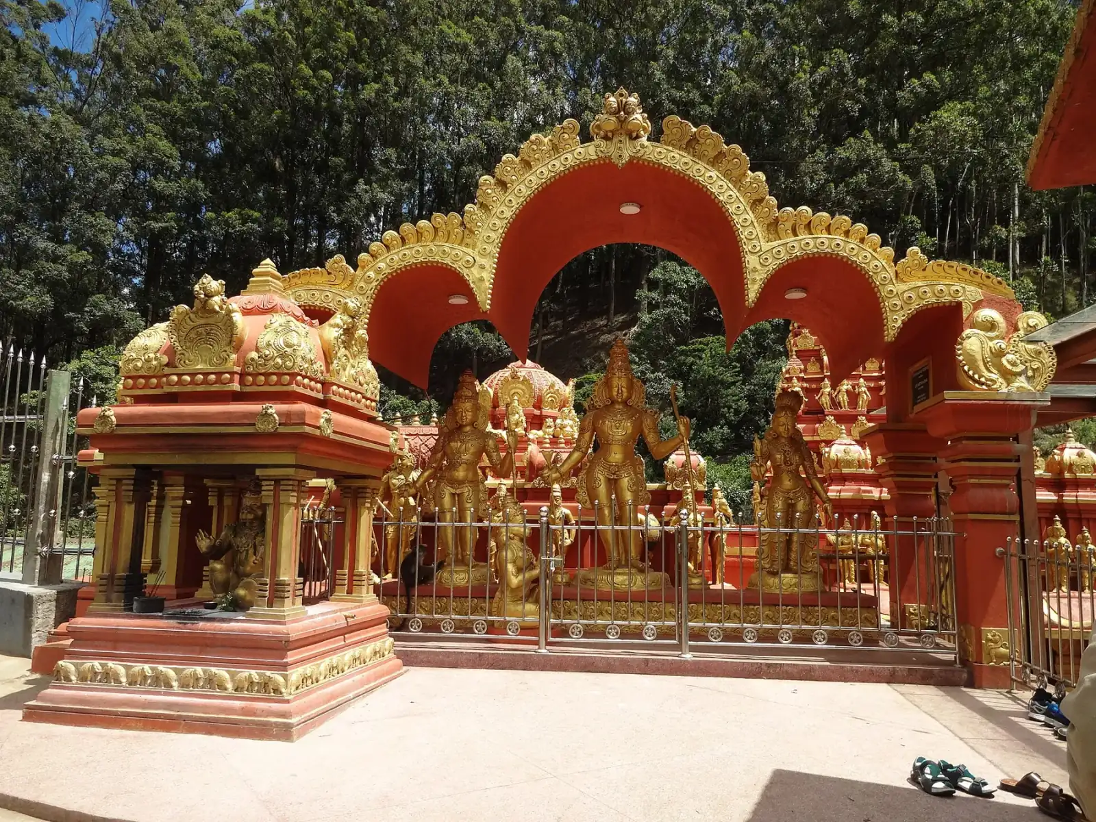 An ornate red and gold arch over golden statues of Hindu deities at the Sita Amman Temple, with forest behind.
