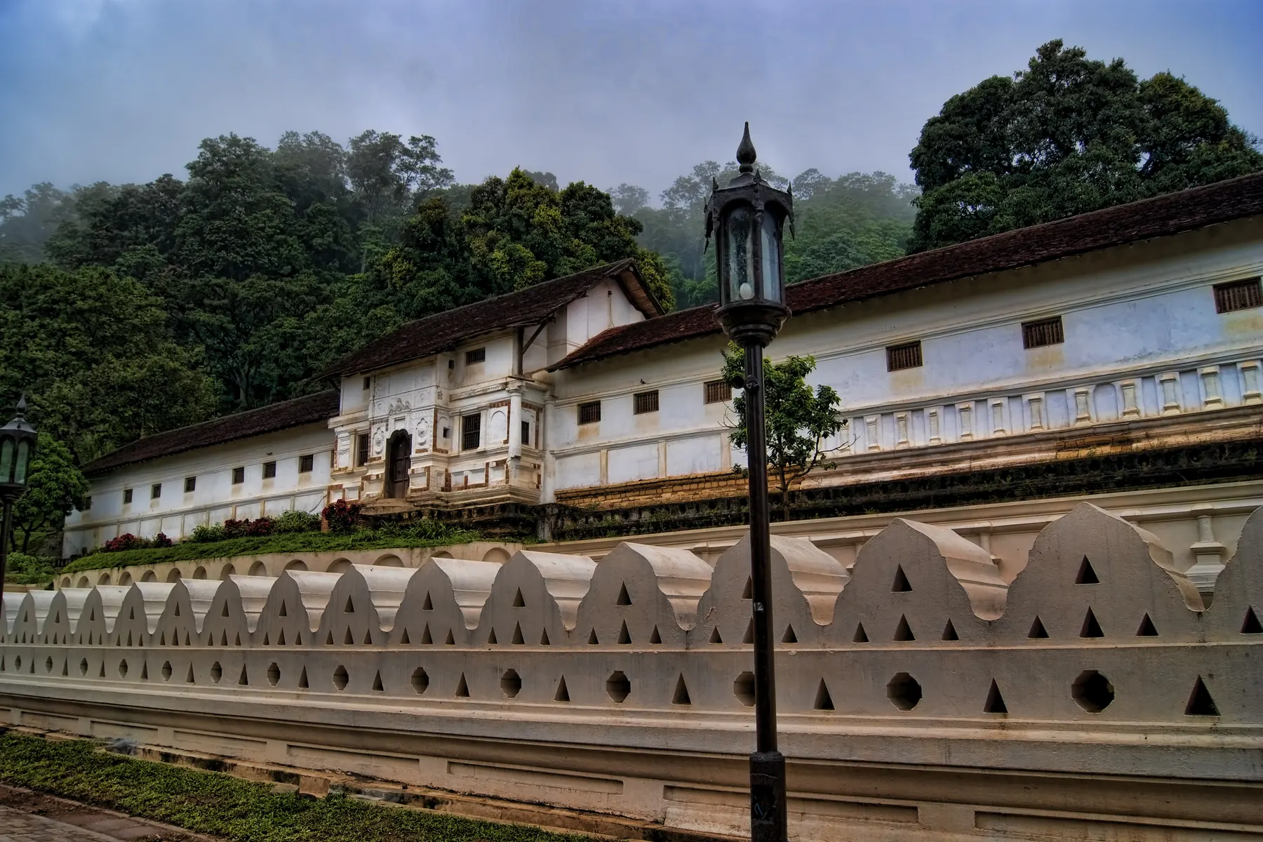 The white walls and tiled roofs of the Temple of the Tooth in Kandy, behind its scalloped wall, with misty hills beyond.