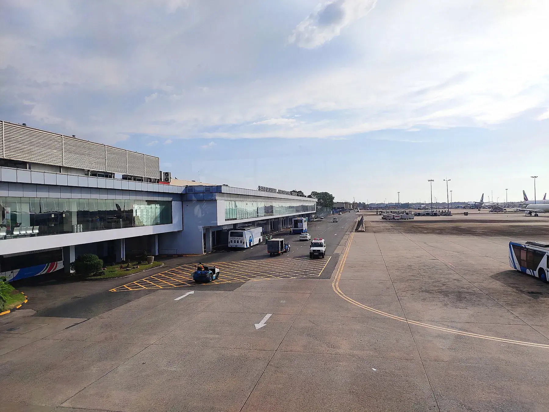The terminal building at Bandaranaike International Airport, with buses and service vehicles on the apron under a cloudy sky.