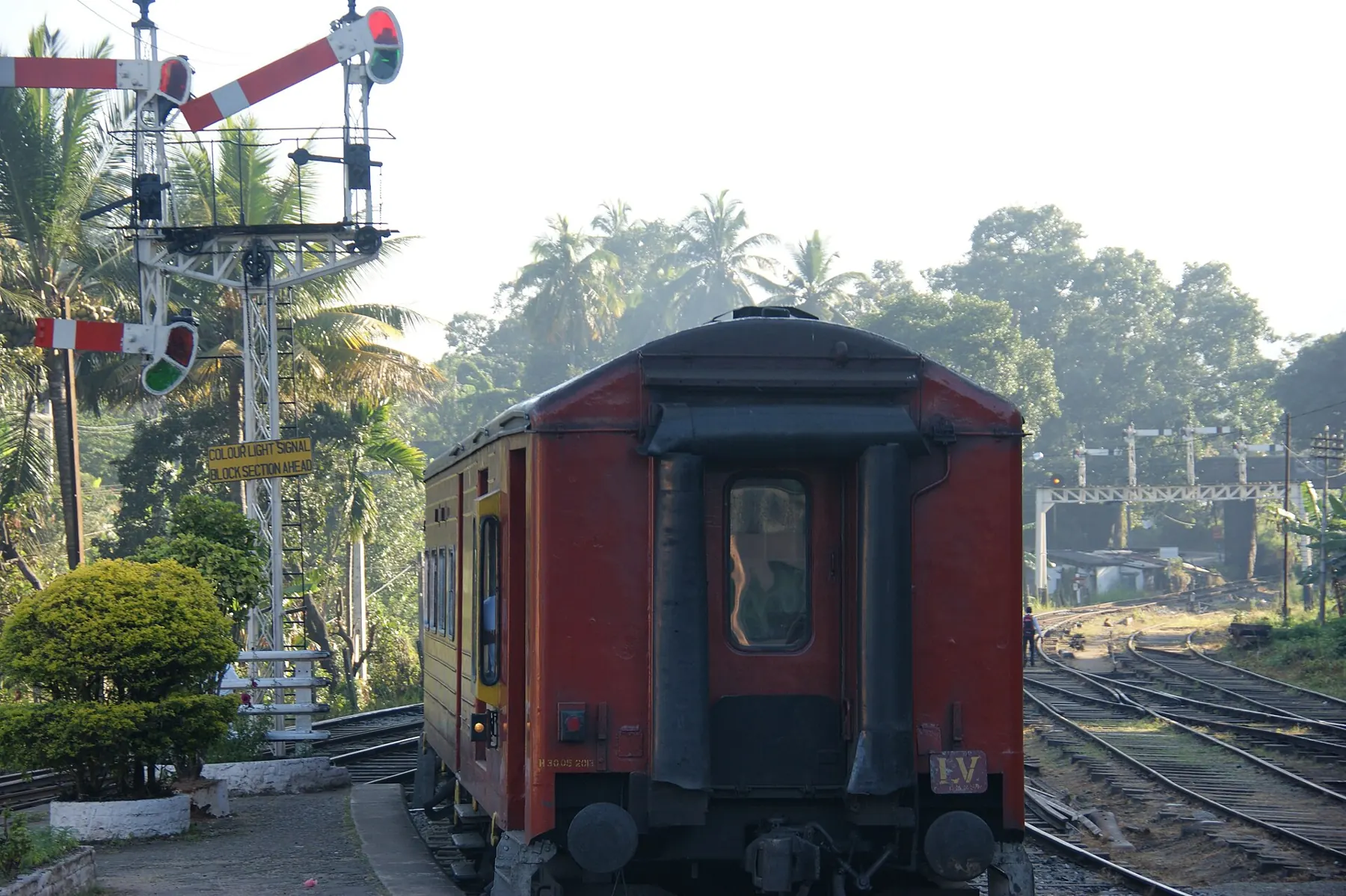 The rear of a red train beside semaphore signals at Peradeniya Junction, framed by palm trees.