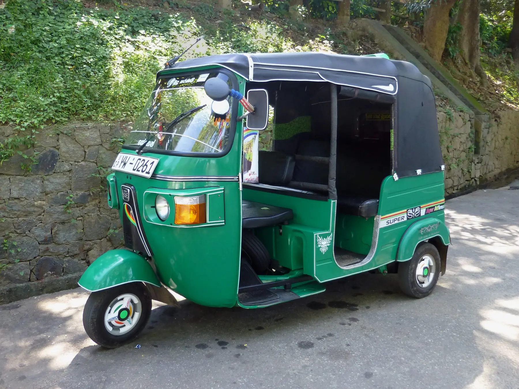 A green three-wheeler tuk-tuk with a black canopy parked on a shaded road beside a stone wall in Kandy.